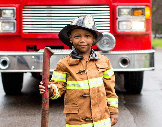 wish kid Tyren dressed in firefighter uniform and hat standing in front of a fire truck