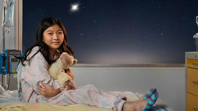 Girl in hospital bed holding a stuffed animal. The night sky is in the background and there is a bright star outside of her room. 