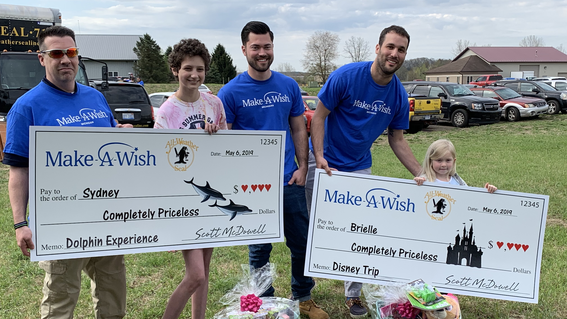 Three adults wearing royal blue t-shirts with the Make-A-Wish Michigan logo pose for a photo with a teenager with short curly brown hair and a child with straight blond hair. Both children hold oversized checks with the Make-A-Wish and All-Weather Seal company logos, symbolizing that All-Weather Seal has adopted their wishes. The checks are made out to the children for a "Completely Priceless" amount, with a description of their wish on the memo line—a dolphin experience for the teenager and a Walt Disney W