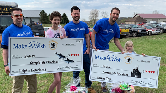 Three adults wearing royal blue t-shirts with the Make-A-Wish Michigan logo pose for a photo with a teenager with short curly brown hair and a child with straight blond hair. Both children hold oversized checks with the Make-A-Wish and All-Weather Seal company logos, symbolizing that All-Weather Seal has adopted their wishes. The checks are made out to the children for a "Completely Priceless" amount, with a description of their wish on the memo line—a dolphin experience for the teenager and a Walt Disney W