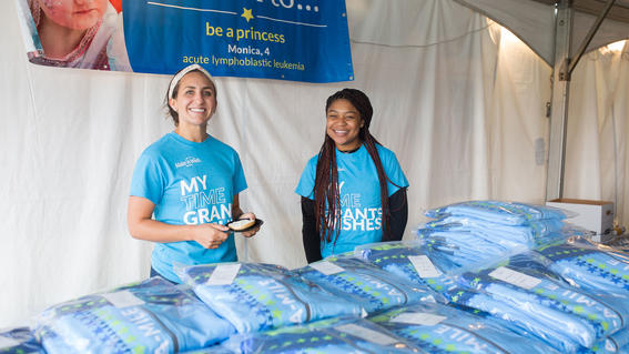 Два молодых взрослых волонтера в бирюзовых футболках стоят за столом, полным поощрительных предметов.
