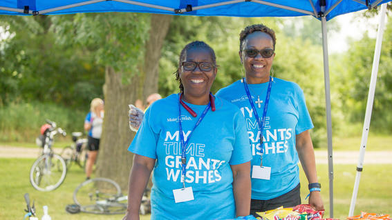 Two adult volunteers wearing turquoise t-shirts, lanyards and reusable gloves stand behind a table full of snacks.