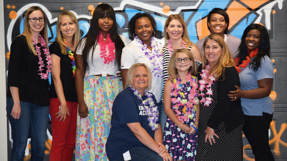 Nine adults in business casual clothing gather around a young child wearing a blue patterned dress and pink lei necklace. The background is a colorful mural.