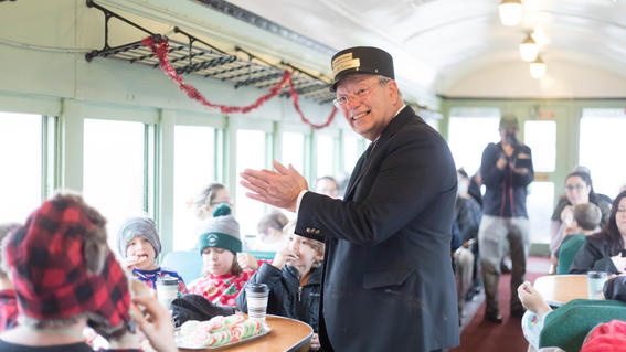 Man dressed as conductor claps hands, leading train full of people in song