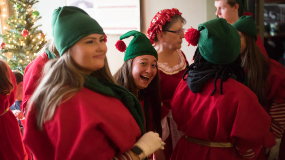 Women dressed as elves stand in a group and smile, a Christmas tree in the background