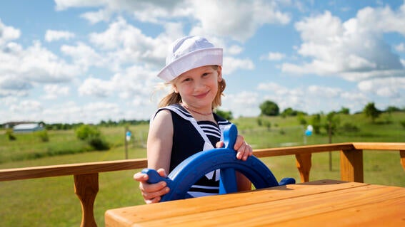 A child wearing a navy and white sailor costume and white sailor hat stands at the captain's wheel of a boat-themed playset.