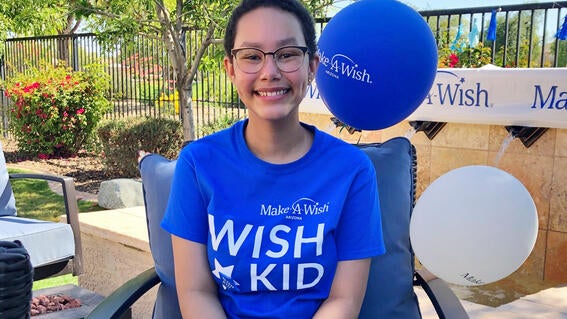 Kylie in blue wish kid shirt, sitting in backyard surrounded by white and blue balloons.