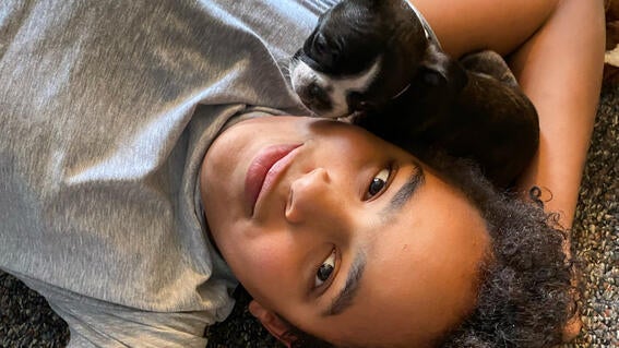 Boy wearing a gray short lying on ground with Boston Terrier puppy by his cheek.