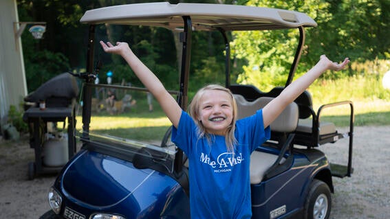 Girl wearing Make-A-Wish shirt holds hands up in excitement in front of blue golf cart.