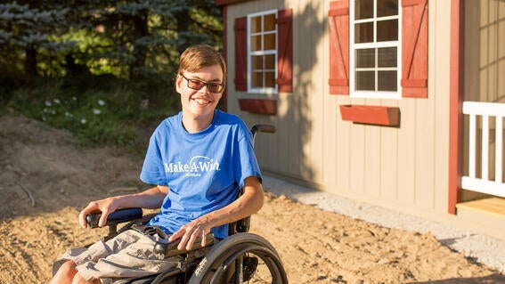 Boy wearing blue Make-A-Wish t-shirt and sunglasses sits in wheelchair outside of his beige outdoor shed with red shutters.