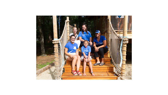 Libby posing on the wooden stairs of her treehouse, wearing her blue Make-A-Wish t-shirt, surrounded by two Make-A-Wish staff members and the two people who are her Make-A-Wish volunteer team