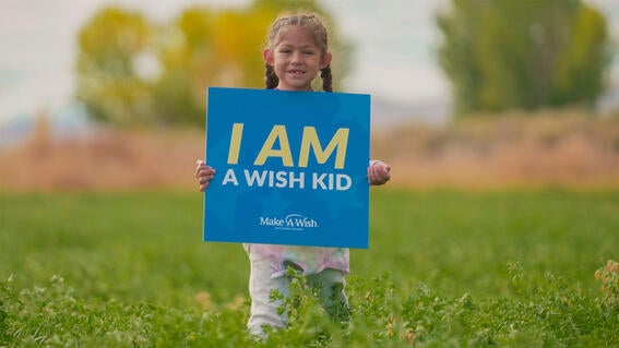 Wish kid Annie smiles in field holding a sign that reads "I am a wish kid"