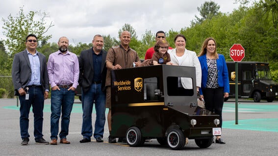 JoJo in his UPS uniform sitting on his mini UPS delivery truck & surrounded by the UPS team