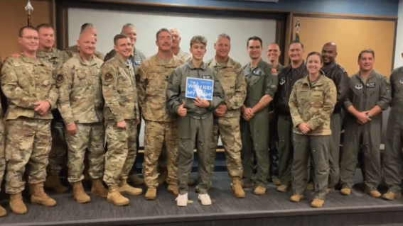 Isaiah holding a sign that says "I'm a wish kid and this is my wish" while standing in the middle of a group of uniformed airforce members