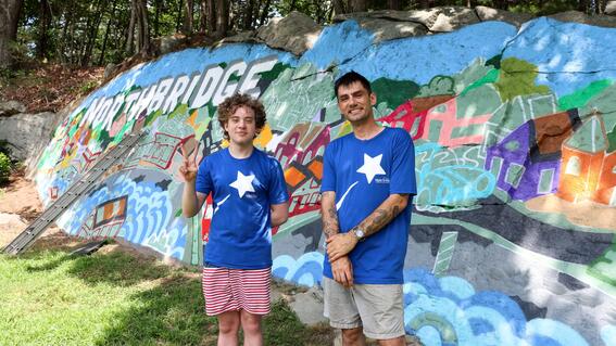 Nate and Ben pose in their Make-A-Wish shirts in front of Ben's colorful mural on a rock face in Northbridge