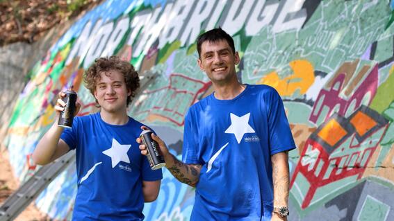 Nate and Ben pose in their Make-A-Wish shirts and cans of spray paint in front of Ben's colorful mural on a rock face in Northbridge