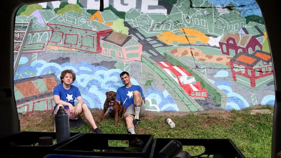 Nate and Ben pose in their Make-A-Wish shirts in front of Ben's colorful mural on a rock face in Northbridge