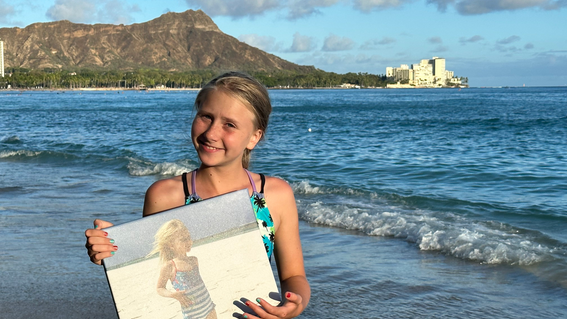 Nora on the beach in Hawaii holding a framed photo of herself on the beach at a younger age
