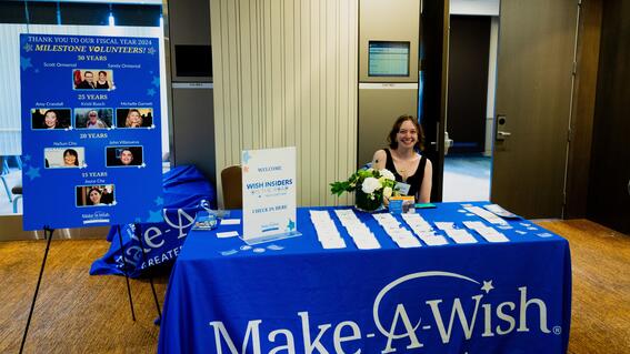 Staff member Rachel Faught sitting at a table with a Make-A-Wish blue table cloth, signing guests in for Wish Insiders