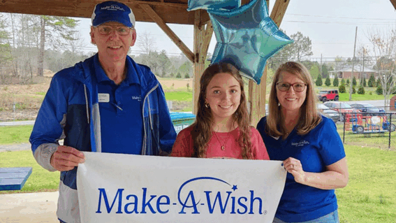 Two volunteers in blue shirts stand on either side of a girl. She holds a sign that says "Make-A-Wish".