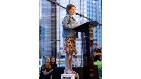 Wish kid Aileen speaks from the podium while standing on a stool, at the 2025 Wine & Wishes fundraising event for Make-A-Wish