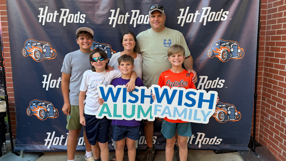 Family of six posing in front of a blue "Hot Rods" backdrop, holding a sign that reads "WISH ALUM FAMILY." The adults are standing in the back, three children in the front.