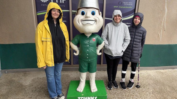 Three people pose with a large statue of a cartoonish statue of the Tin Caps mascot in front of a building; the statue has a smiling face, green shorts, a green top with a logo, a silver bucket for a hat, and stands on a green base labeled "JOHNNY"; the people are wearing jackets, and the person on the far right has a cane and leg braces.
