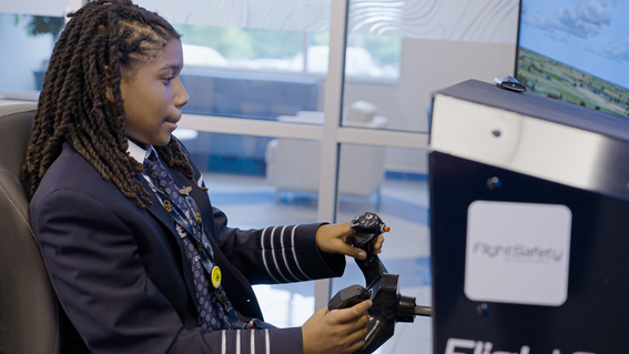 A young Black boy with long, dark lcoks wearing a pilot's uniform, sits in a flight simulator, holding the controls and looking intently at the monitor, with the FlightSafety logo visible on the simulator's body.