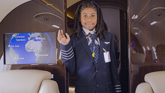 Young Black boy wearing a pilot uniform, smiling and waving, inside a private jet, with a screen displaying a world map showing destination locations.