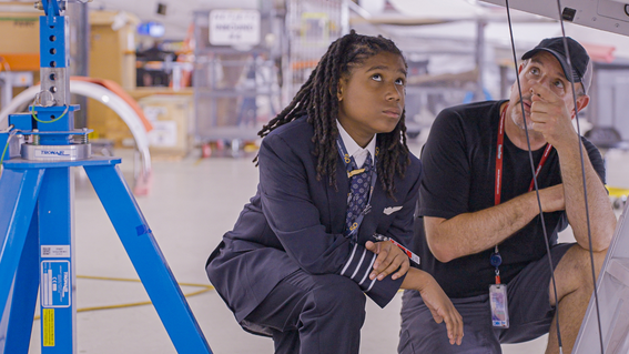 A young boy in a pilot uniform looks up at the underside of an aircraft wing, accompanied by an older man in a cap, both observing the structure in an aircraft hangar.