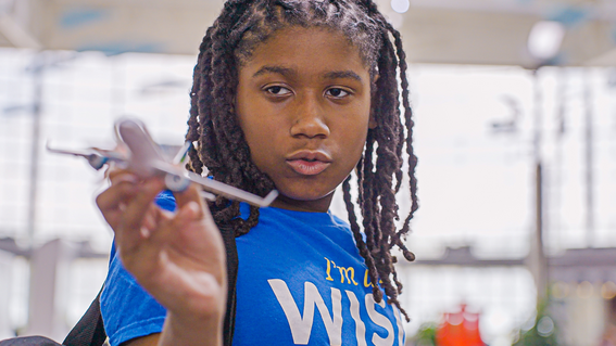 A young Black boy with long, black locks, wearing a blue t-shirt, gazes intensely at a small model airplane held in his hand.  The background appears to be a brightly lit interior space, possibly an airport or educational facility.