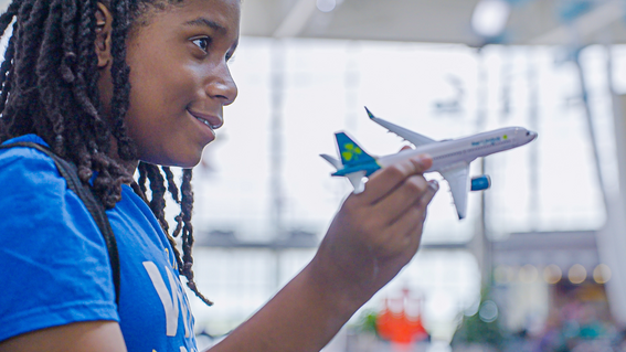 A young person with long, dark locks smiles and holds a toy airplane, in a brightly lit airport setting.