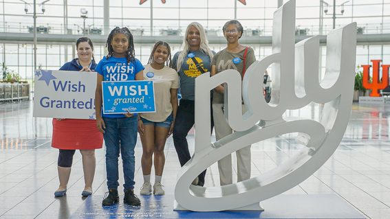 A group of five individuals, including a child wearing a "Wish Kid" shirt, stand with signs reading "Wish Granted!" and pose beside a large, white "Indy" sculpture, celebrating the fulfillment of a wish.