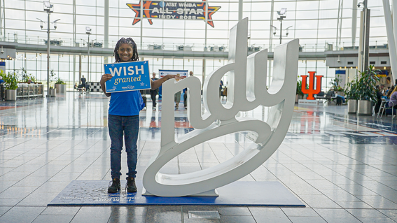 Young person smiling and holding a "Wish Granted" sign in front of a large white "Indy" sculpture, with an "AT&T WNBA All-Star Indy 2025" banner in the background.