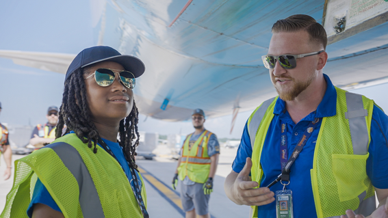 A Black child wearing a baseball cap and sunglasses stands next to a man with a beard and sunglasses, both wearing airport worker safety vests, standing on an airfield under the wing of an airplane, with other workers in the background.