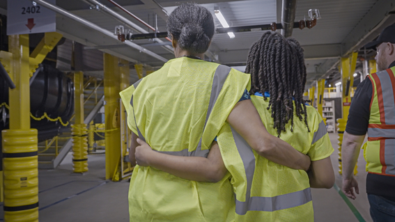 A woman stands next to a child with locks with their arms around one another’s’ waists, wearing yellow reflective vests with a third person walking behind them, wearing a similar vest.