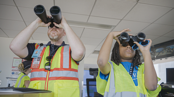 Two people wearing reflective vests look upward, using binoculars. A man with a beard and a nametag reading "Erick," holds large black binoculars in front of him. A child with locks and sunglasses on top of their binoculars stands to the right, also looking upward through binoculars. A third person wearing a reflective vest is partially visible in the background, also looking upward. The setting appears to be an interior office space, with a suspended ceiling and monitors visible in the background.