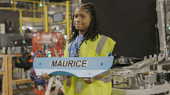 A smiling Black boy wearing a safety vest and a lanyard holds a light blue panel with the name "MAURICE" printed on it in a manufacturing setting, with machinery and equipment visible in the background.