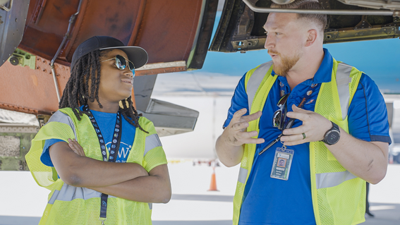 A Black child and a white man stand next to one another and converse under an aircraft wing, the child is wearing sunglasses and the man has a beard,  both in blue shirts and yellow safety vests, converse under an aircraft wing.
