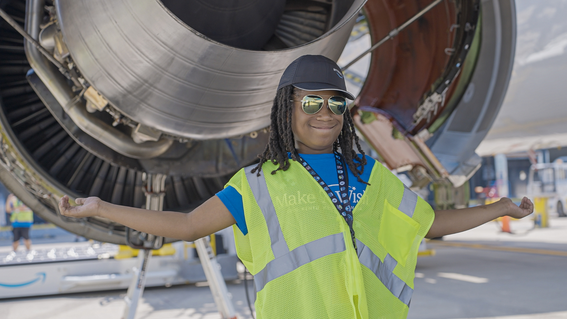 A smiling Black boy with long locks, wearing sunglasses, a black baseball cap, a blue shirt, and a reflective yellow vest, stands in front of a large jet engine with open access panels, arms outstretched. The engine is gray and exposed, and the background shows the edge of an airplane wing and other ground support equipment.