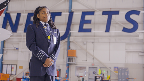Young Black boy in a pilot uniform stands with hands clasped, smiling, in front of a NetJets sign inside a hangar.