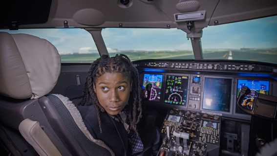 A young Black boy with long locks sits in the cockpit of a simulated airplane, looking at the viewer. He is wearing a dark blazer and is seated to the left of the cockpit controls, with the runway visible through the windshield. The cockpit panel is filled with various displays, buttons, and switches.