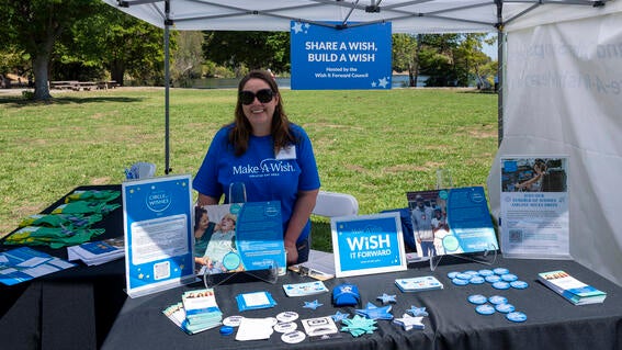 A volunteer in a blue t-shirt stands behind a table with stickers, buttons, and display plaques