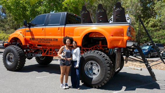 Two women and a small dog stand in front of a large orange monster truck