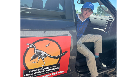 Ian wearing a dinosaur baseball cap and getting out of an official car at the Hell Creek Dino Museum