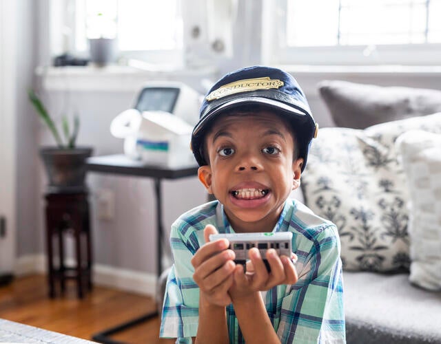 Wish kid Hazael sitting in living room holding toy train with conductor's hat on head