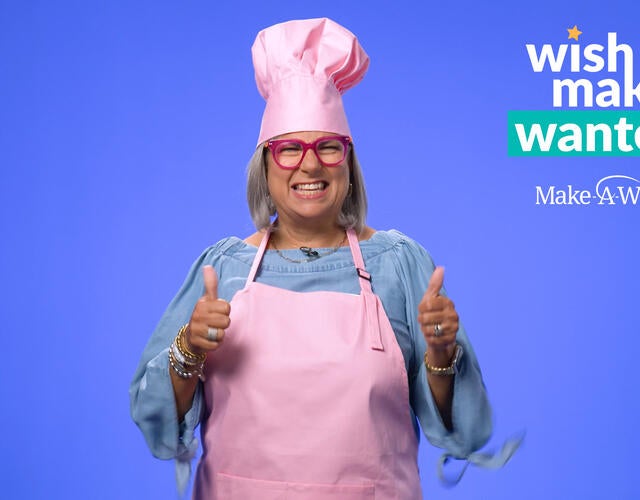 Image of Cathy, a donor, in a pink apron and chef's hat with the Wishmakers Wanted logo in the top right corner
