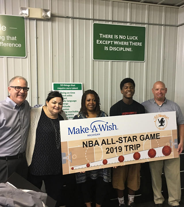 Four adults stand with a teenage wish kid in front of a wall that has many signs with inspirational sayings. The group of people holds a large countdown sign with the Make-A-Wish Michigan and All Weather Seal company logos and images of basketballs and a basketball court.