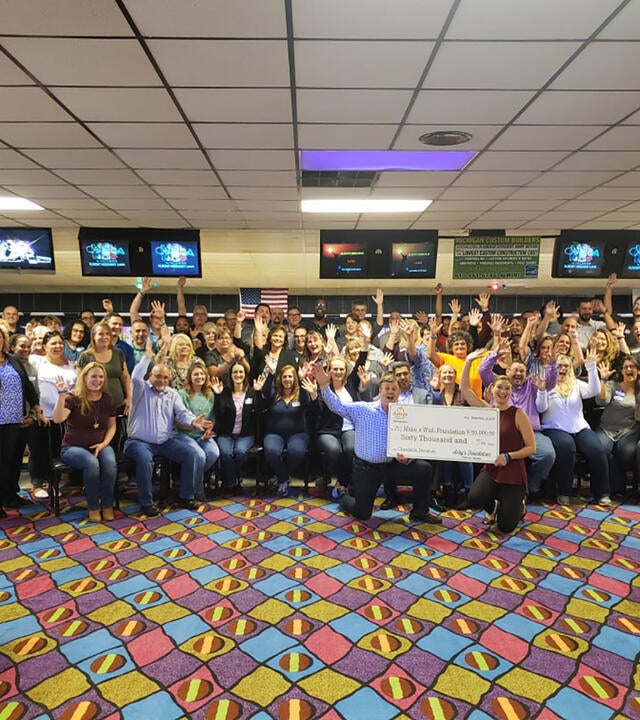 A large group of people gather together in a bowling alley for a photo. Many of them have thrown their hands in the air. In the front of the group, two adults are kneeling and holding an oversize check, made out for sixty thousand dollars to Make-A-Wish Michigan from Arby's.