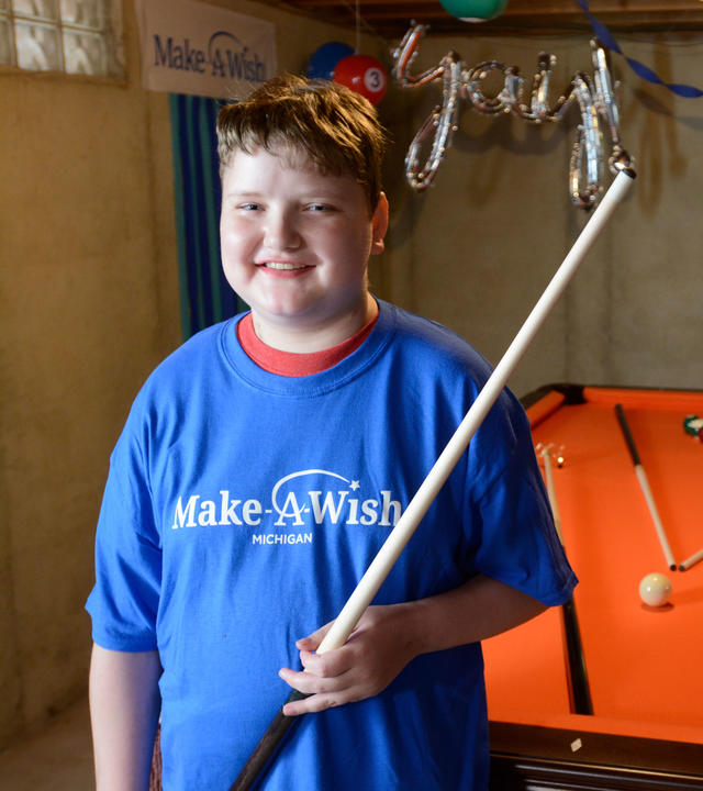 A child with short brown hair wearing a royal blue Make-A-Wish t-shirt stands with a billiard cue in hand in front of a pool table with orange felt. Three additional cues, balls, and other billiards equipments are scattered on the table. In the background above the child's head is a silver balloon that says "yay!"